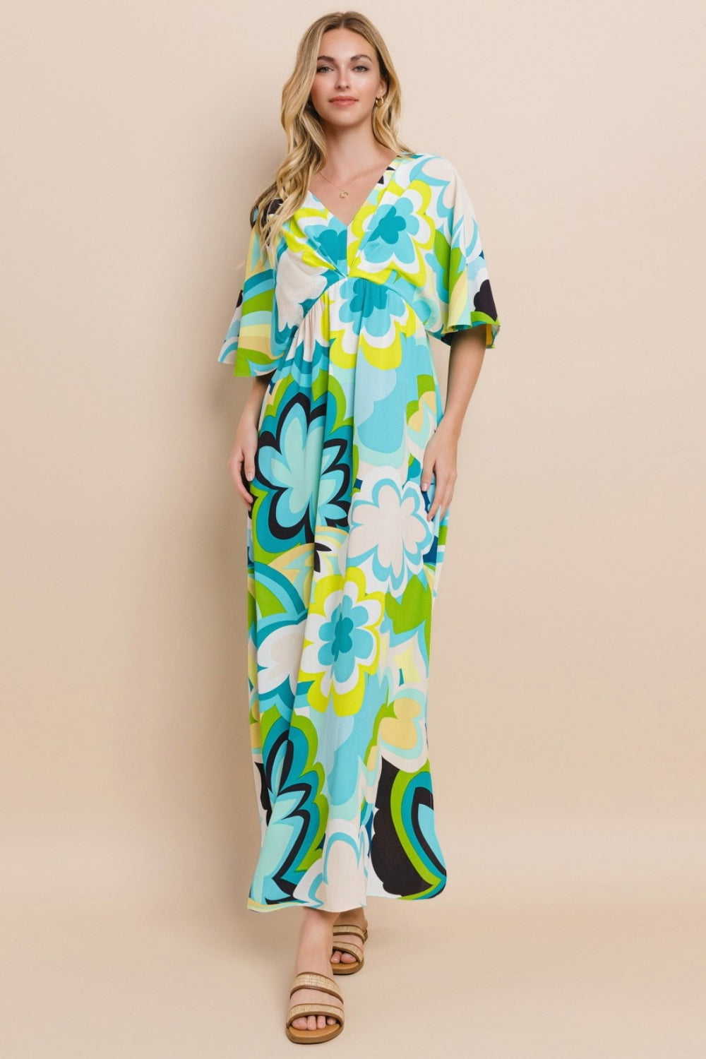 Against a simple backdrop, a woman models the ODDI Floral Printed Slit Maxi Dress, exuding a sense of versatile elegance. This maxi dress embodies summer chic with its vibrant floral design featuring shades of blue, green, and yellow.