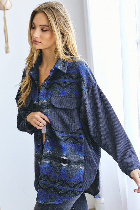 A woman with long blonde hair wears a blue and gray patterned shirt over a white crop top and dark jeans, standing indoors near a window, her casual **Printed Button Down Long Sleeve Jacket** draped casually over her shoulders.