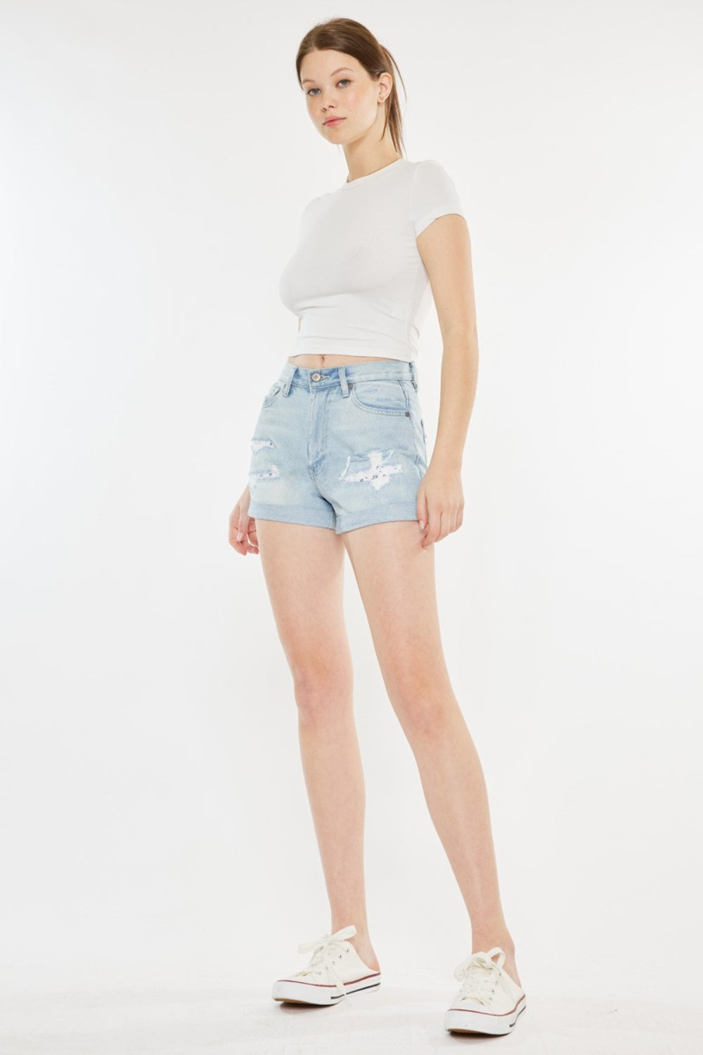 A person stands against a plain background, wearing a white short-sleeve crop top, Kancan High Rise Repaired Mom Denim Shorts in light blue, and white sneakers.