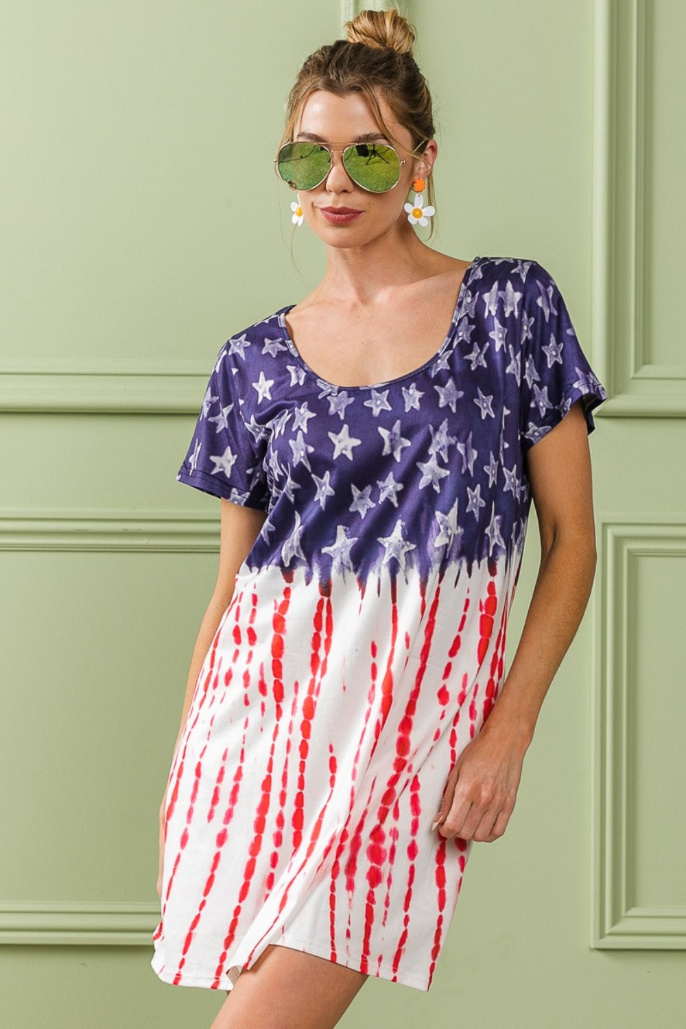 A person wearing the BiBi American Flag Theme Tee Dress, accessorized with sunglasses and earrings, stands against a light green wall with decorative molding.