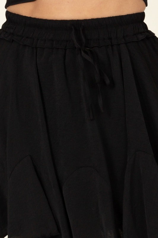 A person is partially visible in the image wearing the Not Your Girl Drawstring Ruffled Mini Skirt, featuring a bright pink flared silhouette with an elastic waistband and drawstring, paired with a matching top.