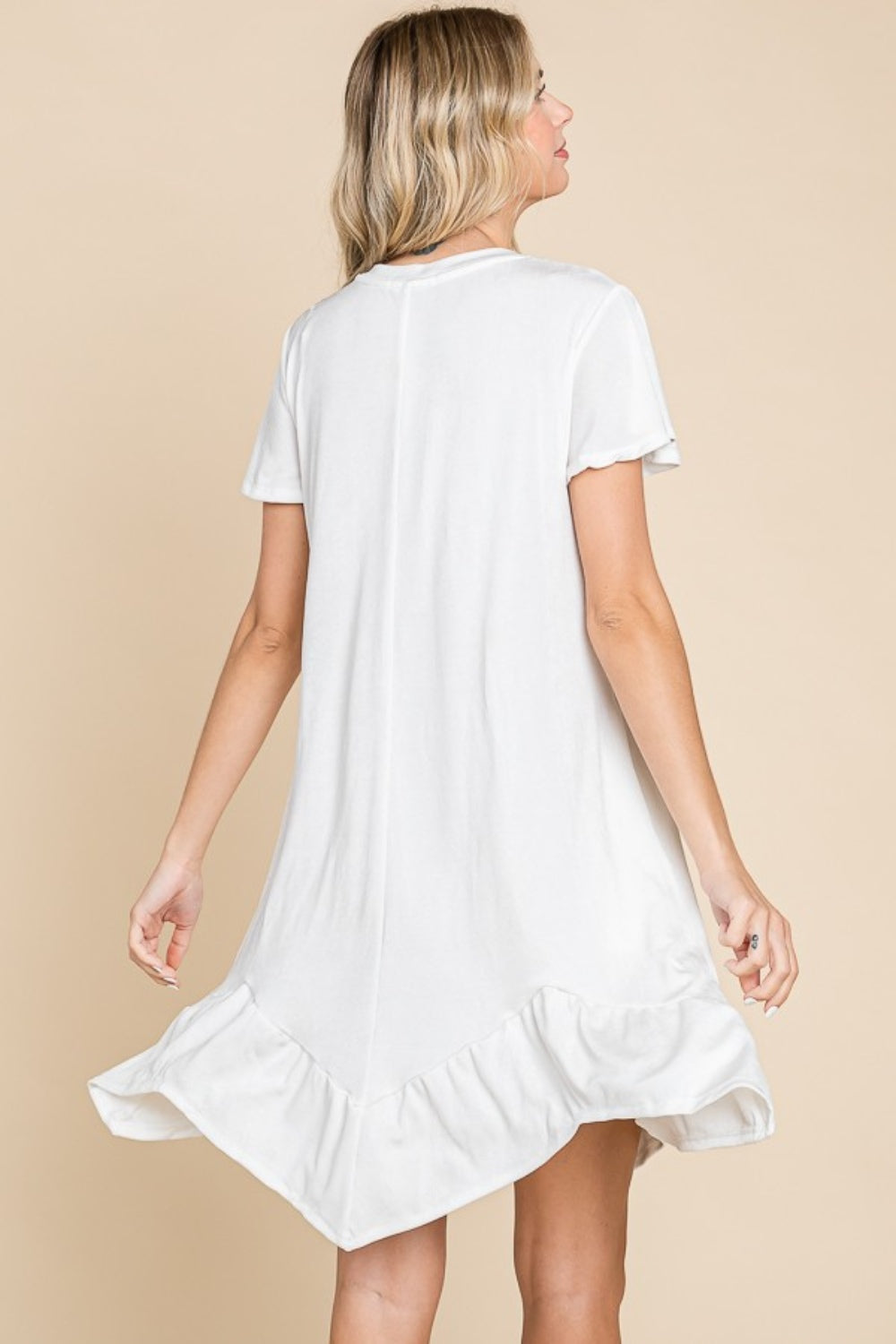 A person wearing the versatile white Culture Code Full Size Short Sleeve Ruffled Asymmetric Hem Dress stands in front of a plain beige background, showcasing its flattering fit.