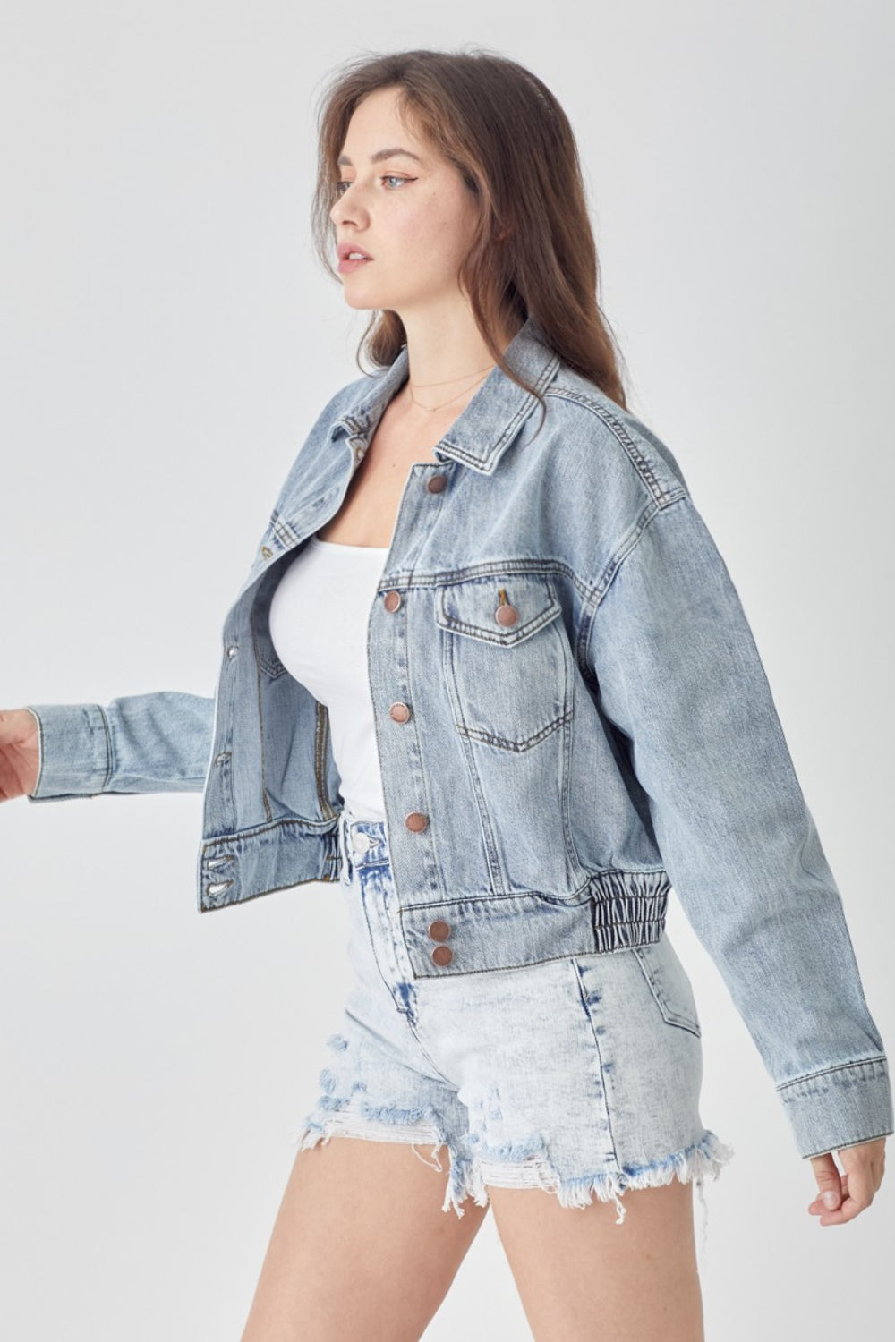 A woman wearing the RISEN Full Size Button Down Cropped Denim Jacket over a white top and distressed denim shorts stands against a plain background, showcasing versatile outerwear perfect for any casual occasion.