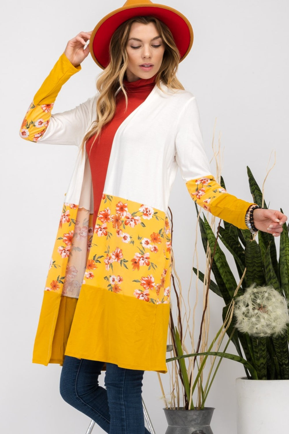 Wearing the Celeste Full Size Floral Color Block Open Front Cardigan with a white and yellow floral print, along with a red hat, a red top, and jeans, a woman stands beside a houseplant.