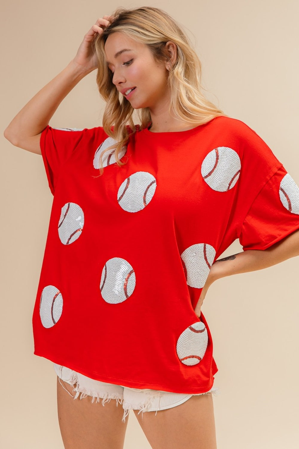 Woman wearing the BiBi Sequin Baseball Drop Shoulder Short Sleeve T-Shirt in red, paired with white shorts, posing against a beige background.