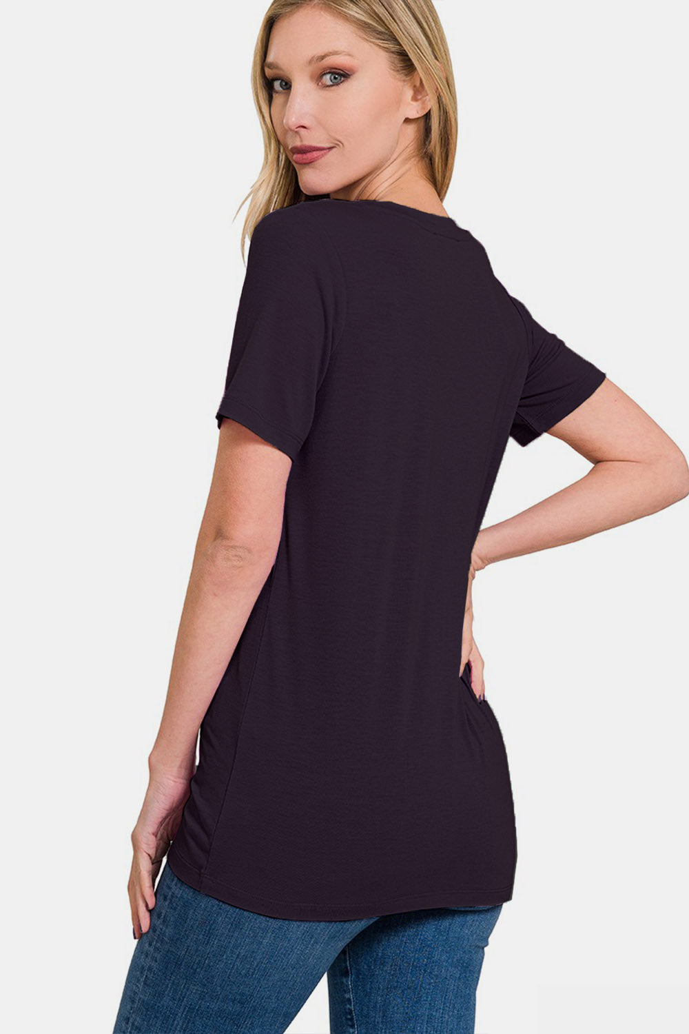 A woman with blonde hair is wearing the Zenana V-Neck Short Sleeve T-Shirt in black, made of breathable fabric, paired with blue jeans. She is standing against a plain white background, smiling. Perfect for everyday wear.