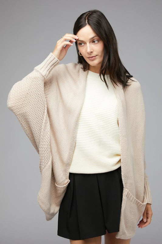 A person stands against a plain background, wearing a Pattern Knit Dolman Sleeve Solid Slouch Cardigan in textured blue over a white sweater with distressed white shorts, hands in cardigan pockets.