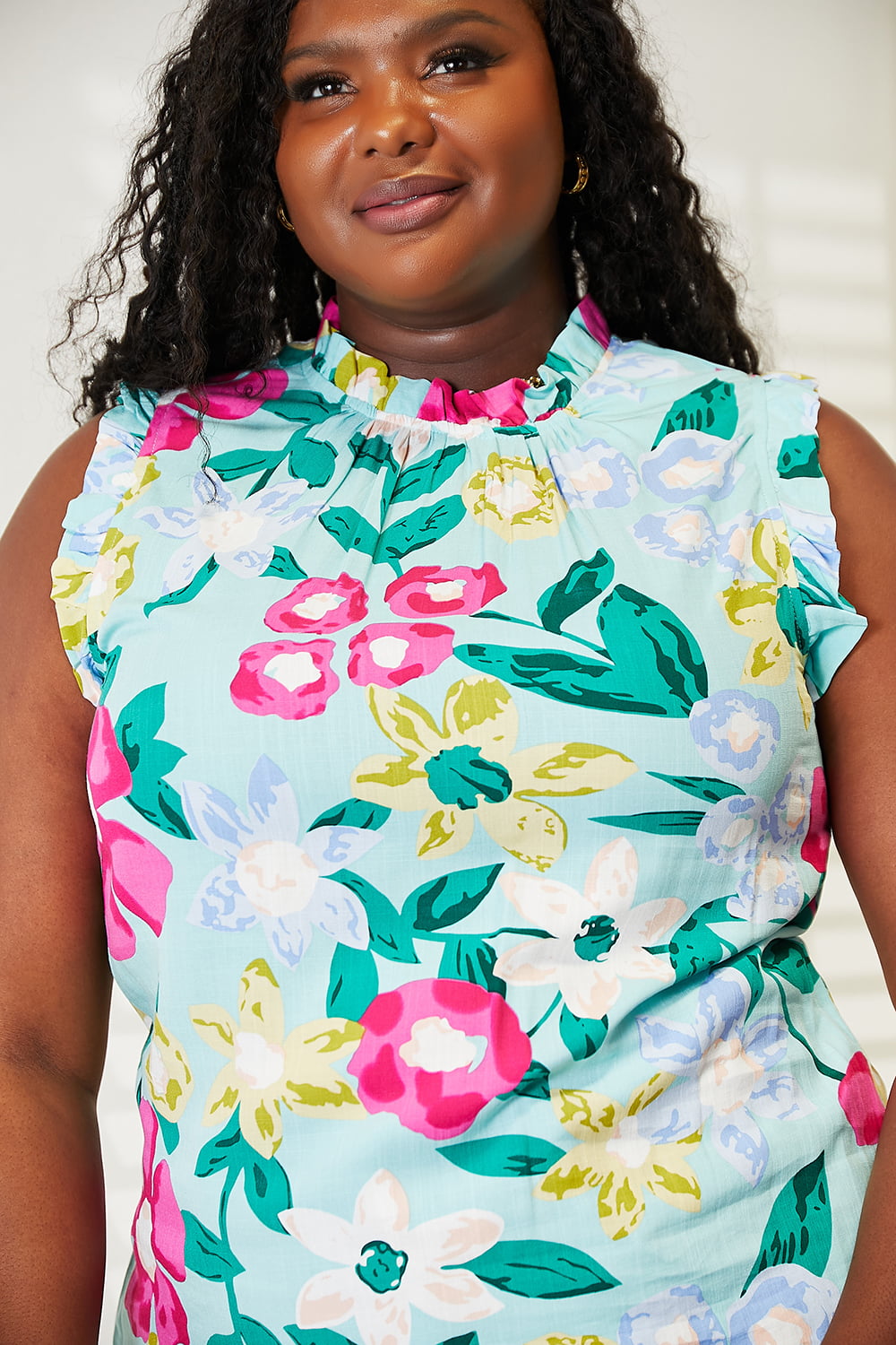 A person wearing the Double Take Floral Print Ruffle Shoulder Blouse and jeans stands indoors, leaning against a piece of furniture, while smiling at the camera.