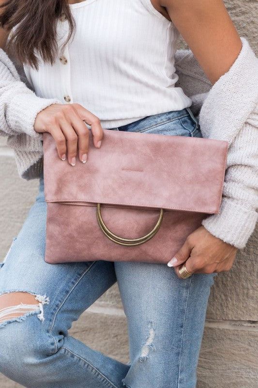 A person holds a tan Fold-over O-ring Clutch with a circular handle in front of their floral skirt and cream top.