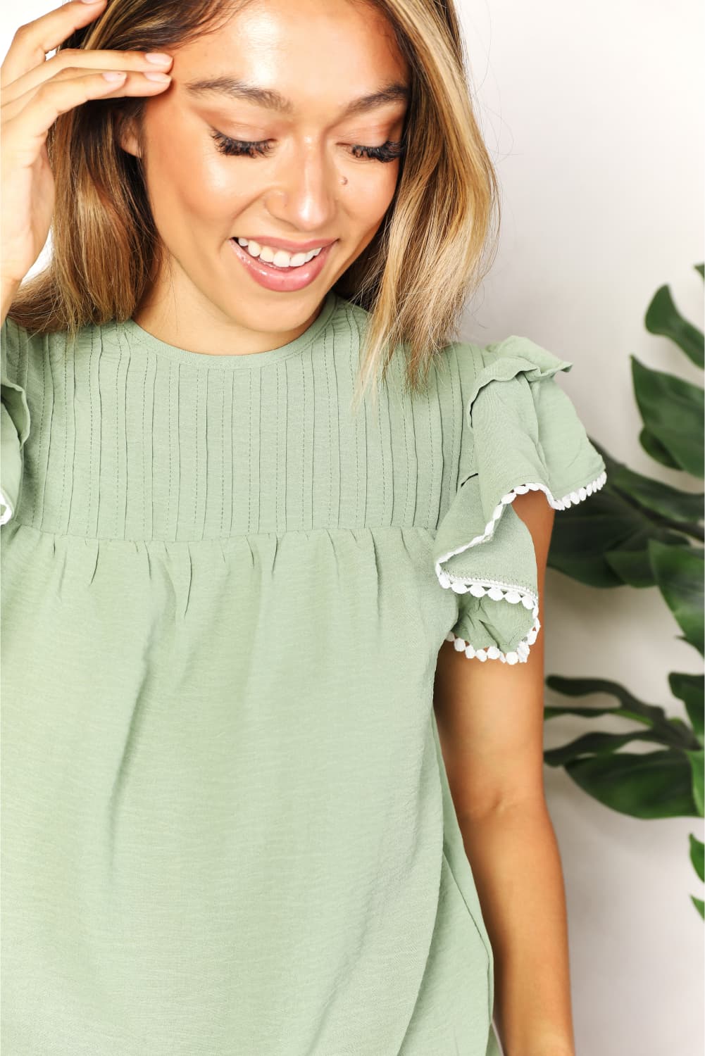 A woman stands and smiles, wearing the Perfee Pleated Detail Flutter Sleeve Blouse in light green paired with black pants.