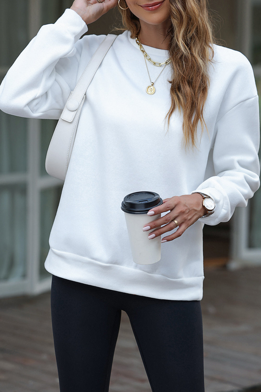 A person wearing a white beanie, the EVERY THING WILL BE OKAY Colorful Letters Sweatshirt, and sunglasses, holding a coffee cup, stands outside near windows.