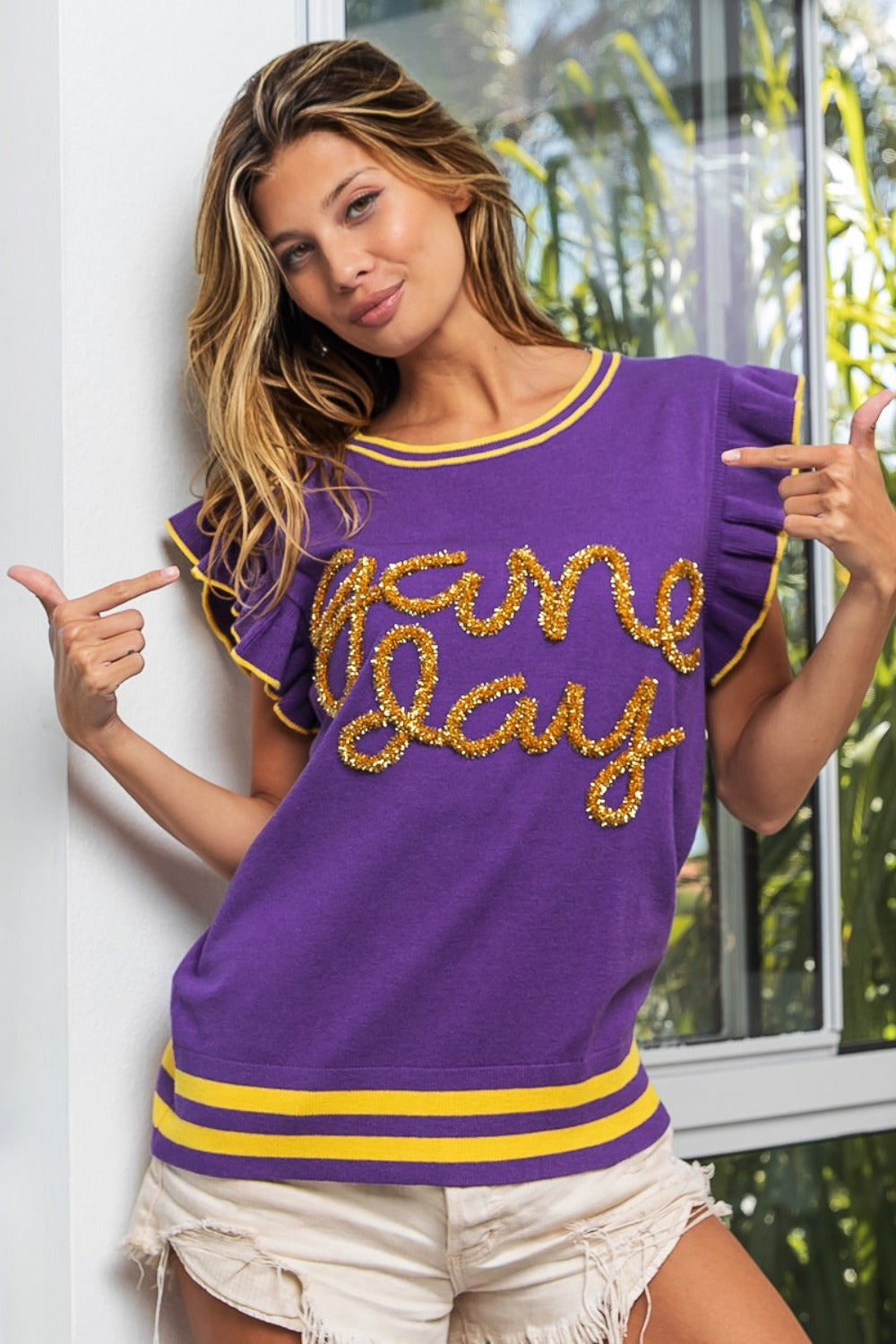 A person wearing the BiBi Game Day Letter Contrast Trim Ruffled Sleeveless Sweater, with its purple color and gold "Game Day" lettering, gestures towards the text. They stand by a window, pairing it with white shorts that accentuate their lively ensemble's contrast trim.