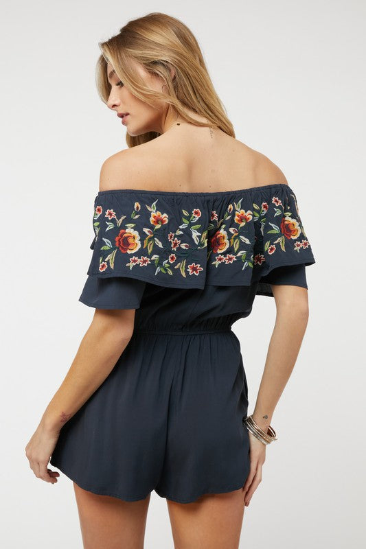 Wearing the stylish Floral Embroidered Off Shoulder Romper, a woman poses gracefully, showcasing its ruffle detail and making it an ideal addition to any summer wardrobe.