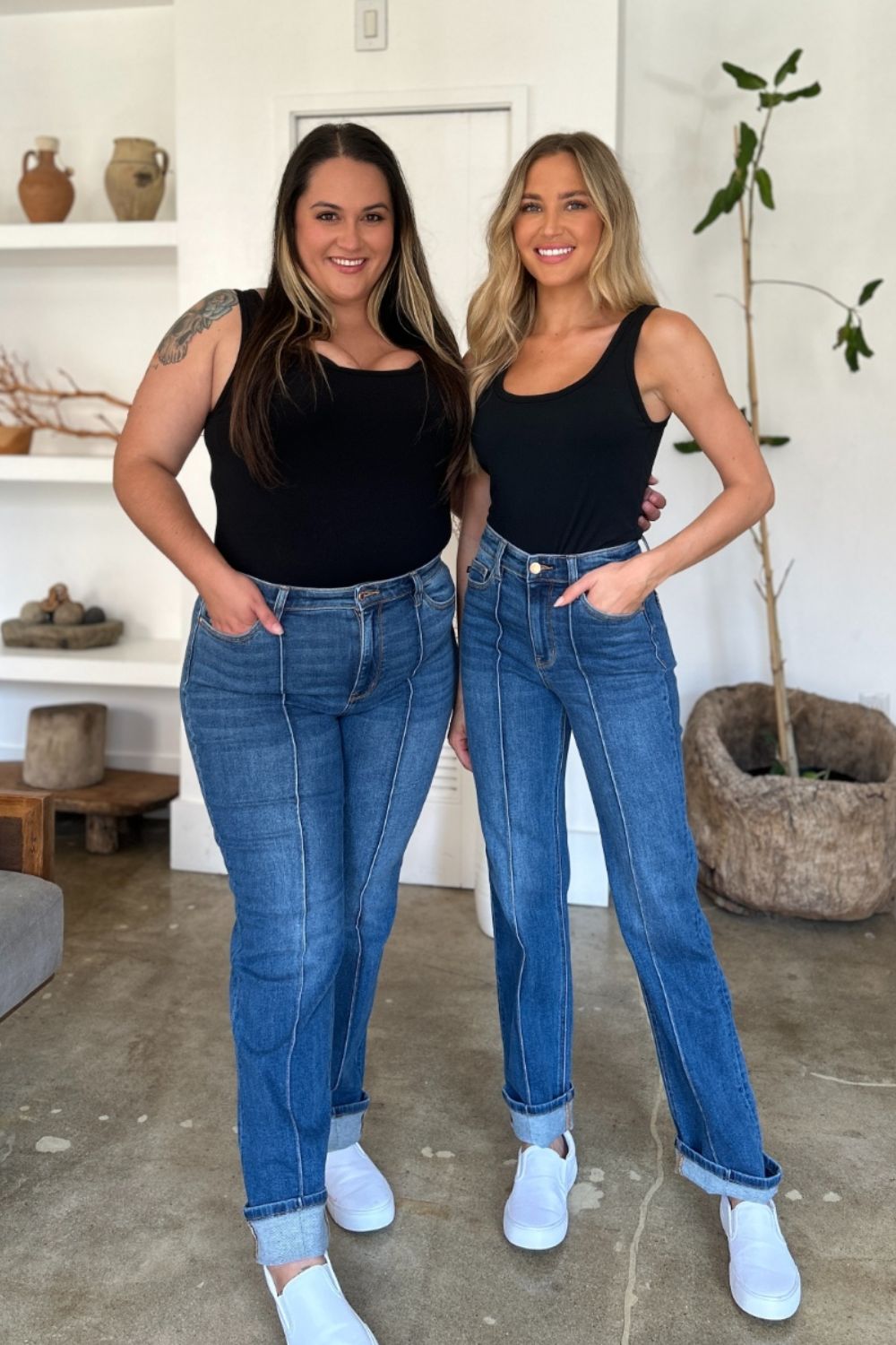 Two women stand side by side indoors, both wearing black tank tops, Judy Blue Full Size High Waist Front Seam Detail Straight Jeans, and white shoes. They are smiling and posing for the camera. A plant and shelves with decorations are in the background.