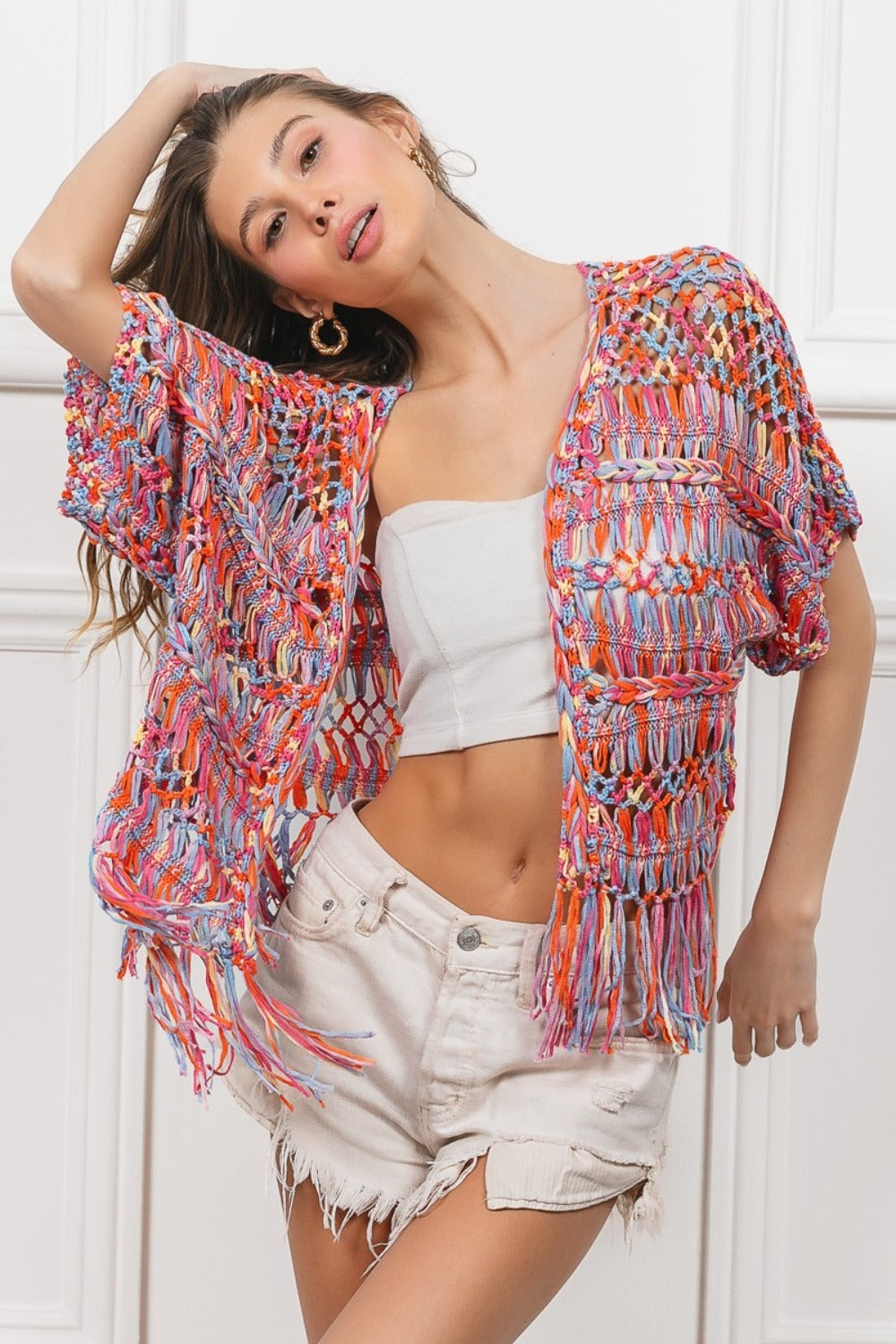 A woman wearing a BiBi Open Front Fringed Crop Knit Cardigan, a white bandeau, distressed white shorts, and a wide-brimmed straw hat stands against a white paneled background, smiling with one hand on her hat.