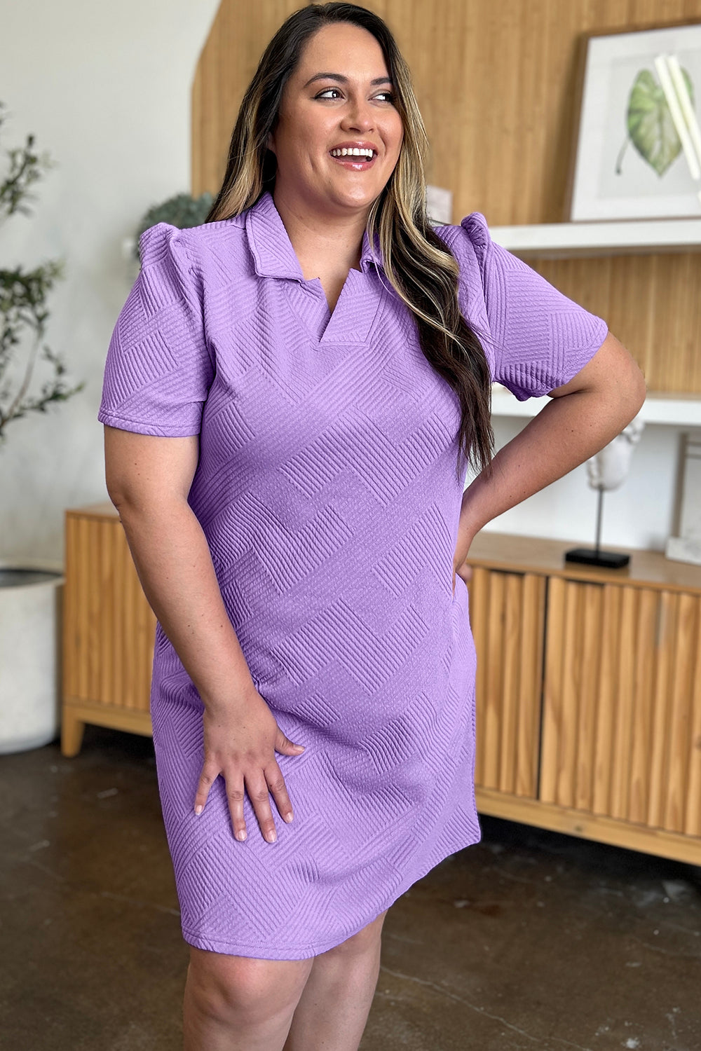 A woman wearing the Double Take Full Size Texture Collared Neck Short Sleeve Dress, featuring a geometric pattern in purple and made from soft polyester, stands against a neutral background. She has long, light hair and is looking slightly to the side. The dress is available in sizes S-3XL and is easy to care for—just machine wash cold.