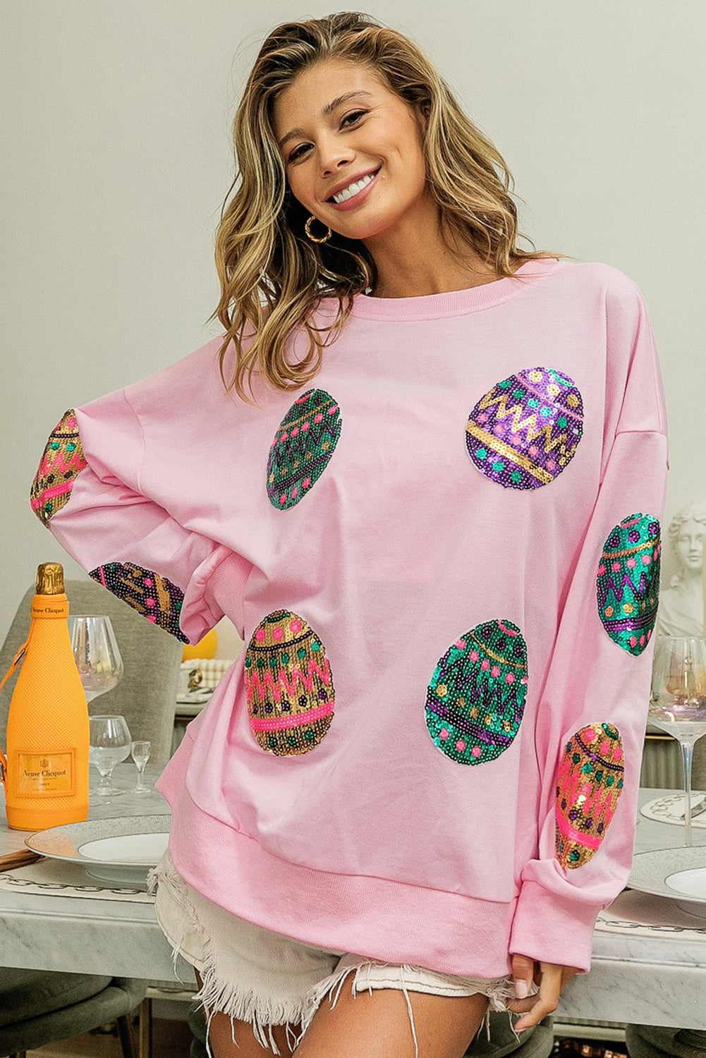A woman with long, wavy hair wears a Pink Sequined Easter Egg Drop Shoulder Oversized Sweatshirt featuring colorful, patterned circles. She is seated in a dining area with a table set with glasses, plates, and two bottles, one of which is orange.