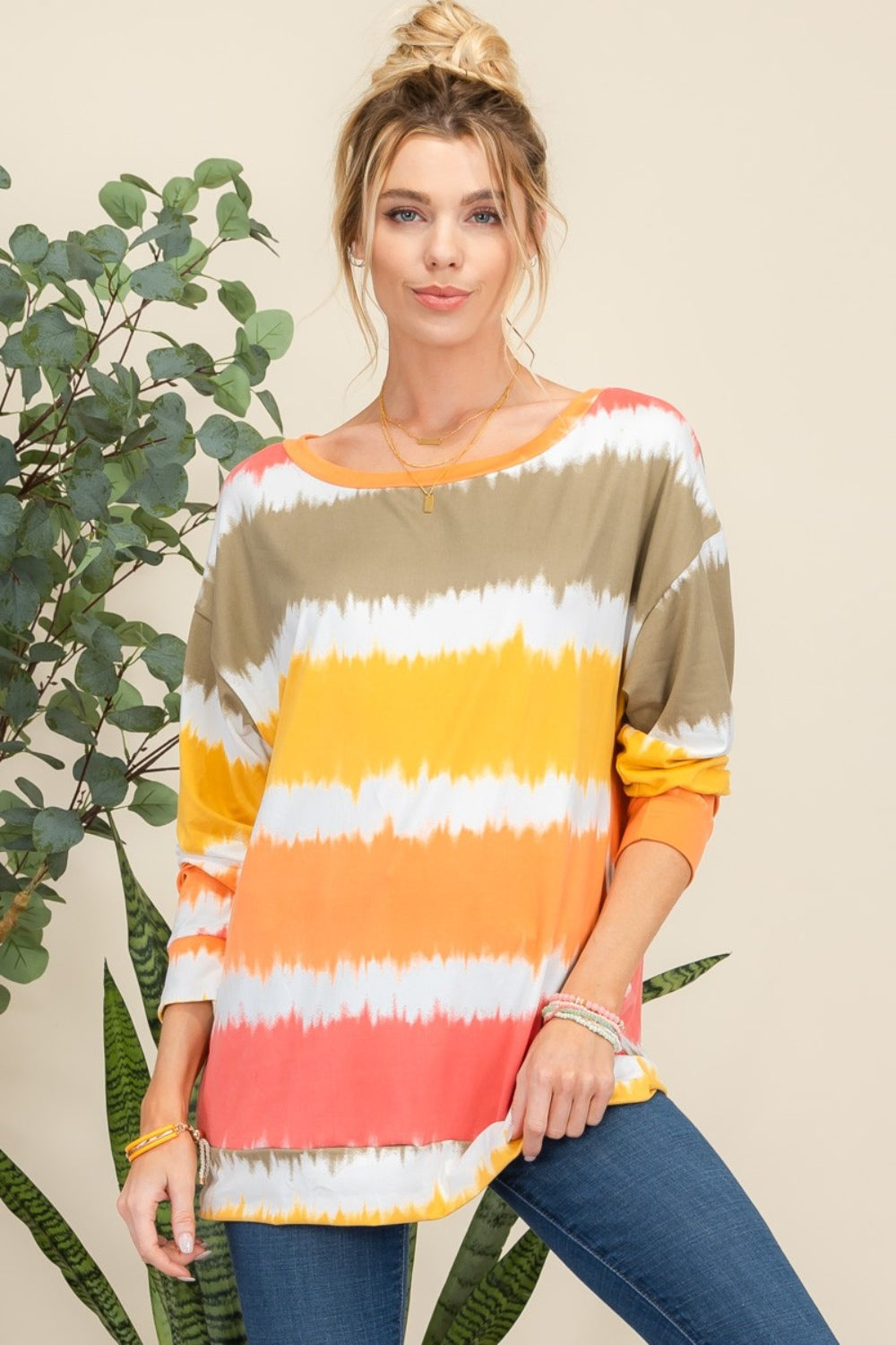 Wearing the Celeste Full Size Striped Long Sleeve T-Shirt paired with jeans, a person smiles while standing against a light background adorned with plants, exuding a casual yet stylish look.