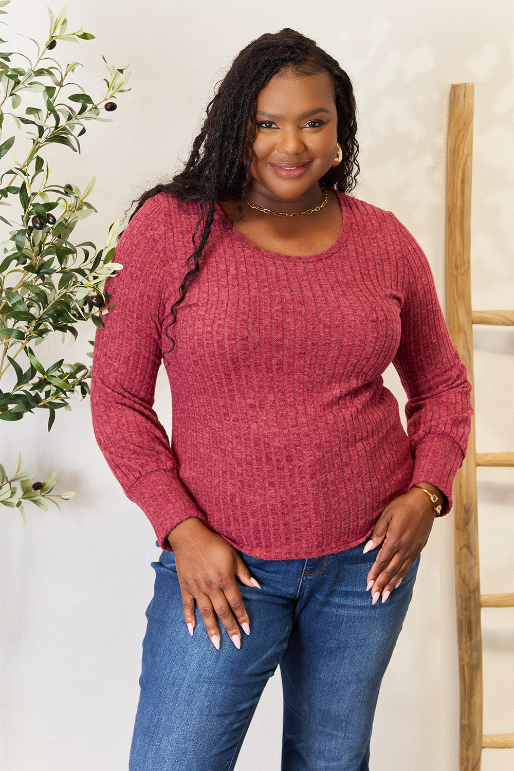 A person in a stylish maroon Ribbed Round Neck Lantern Sleeve Blouse and jeans stands by a wooden ladder, with olive branches in the background.