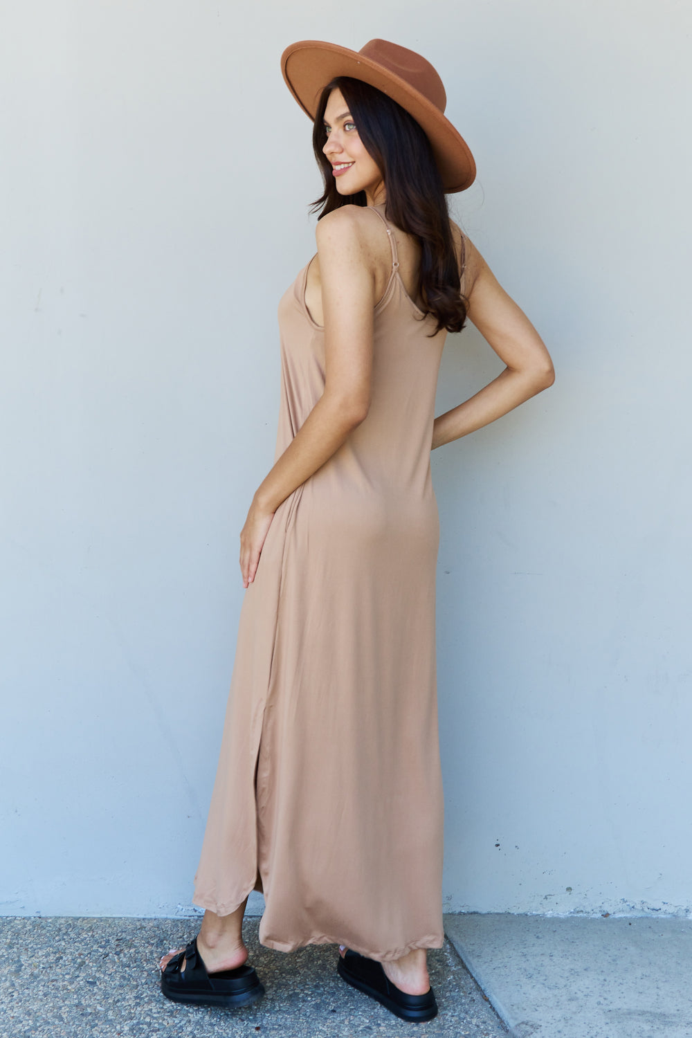 A woman stands against a plain background, wearing the Ninexis Good Energy Full Size Cami Side Slit Maxi Dress in Camel along with a wide-brimmed hat. She has one hand on her hip and is smiling.