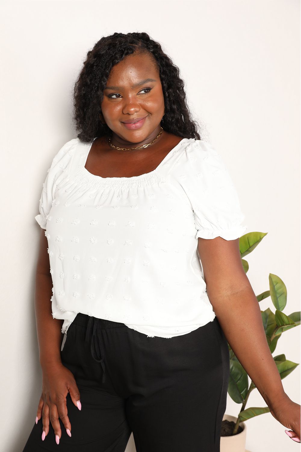 A person wearing the Mandy Swiss Dot Puff Sleeve Square Neck Blouse in white, paired with black pants, stands in front of a light background with green leaves visible on the right side.