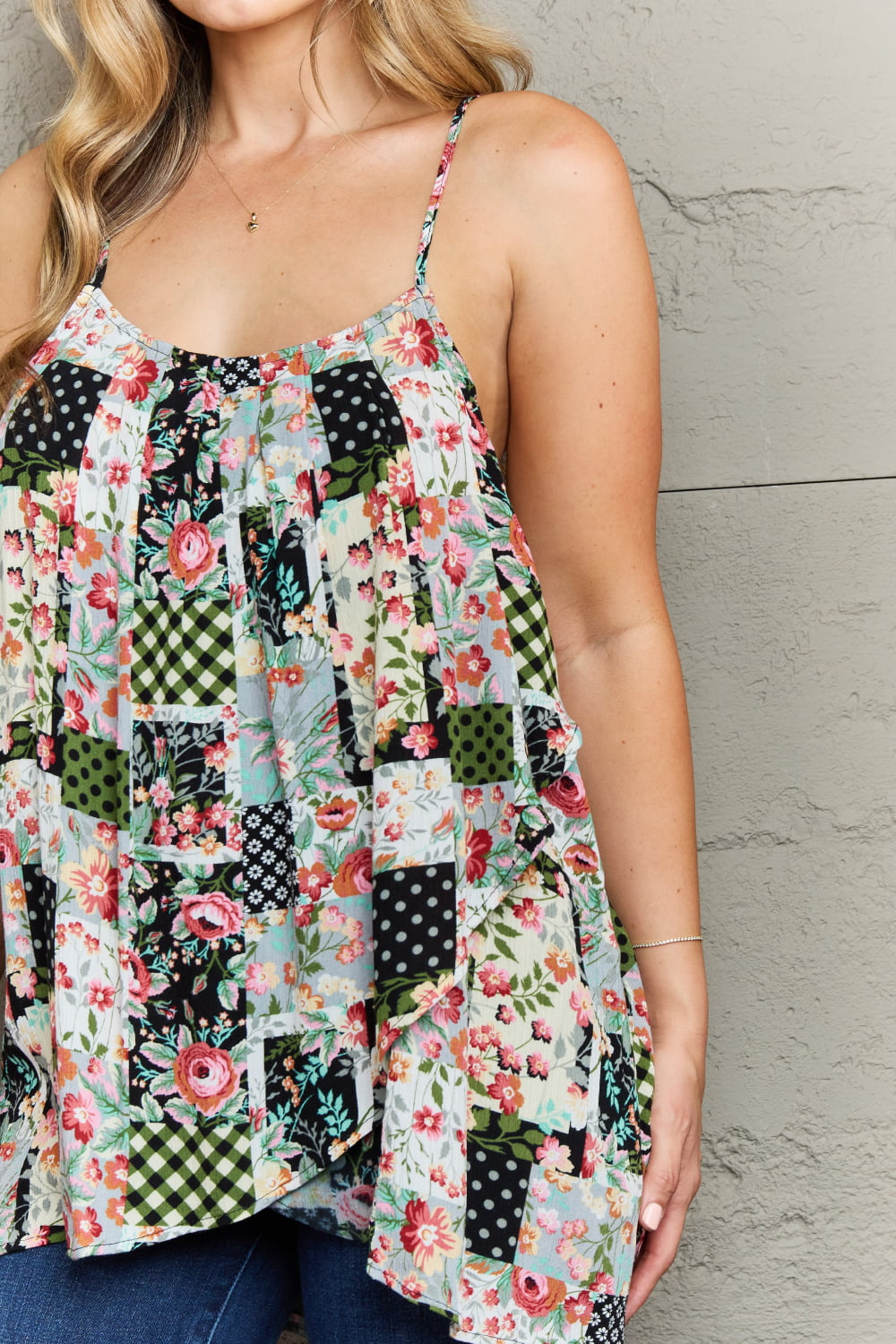 A woman wearing a Ninexis Hang Loose Tulip Hem Cami Top with a stylish floral and checkered pattern stands in front of a textured gray wall, her hands resting by her sides and a relaxed expression on her face.