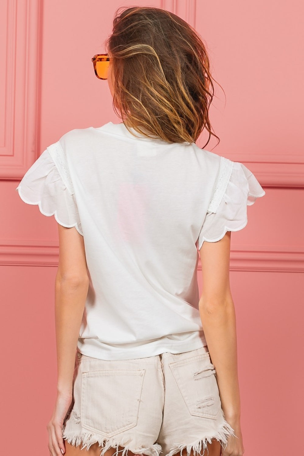 A woman with wavy hair dons the elegant BiBi Scallop Edged Lace Trim Top and large amber sunglasses while standing in front of a vibrant pink wall.