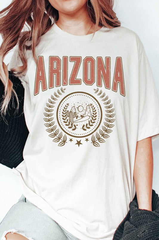 A person wearing a light gray ARIZONA Graphic Tee that showcases a desert scene with cacti and a sun, encircled by a laurel wreath design. This tee is made from 100% cotton and is available in unisex sizing.