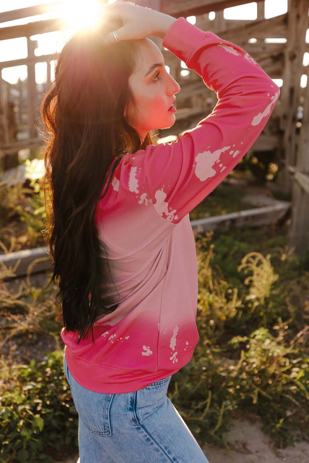 A person with long dark hair is standing outdoors, facing away from the camera, wearing a Pink Mama Rhinestone Letter Graphic Tie Dye Pullover and light blue jeans. Old wooden structures are visible in the background.