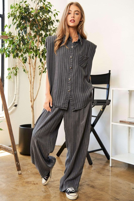 A person wears a casual outfit called the Stripe Button Down Shirt and Long Pants Set, which features a loose-fitting pinstriped design, paired with sneakers. They stand indoors near a potted plant and a director's chair.