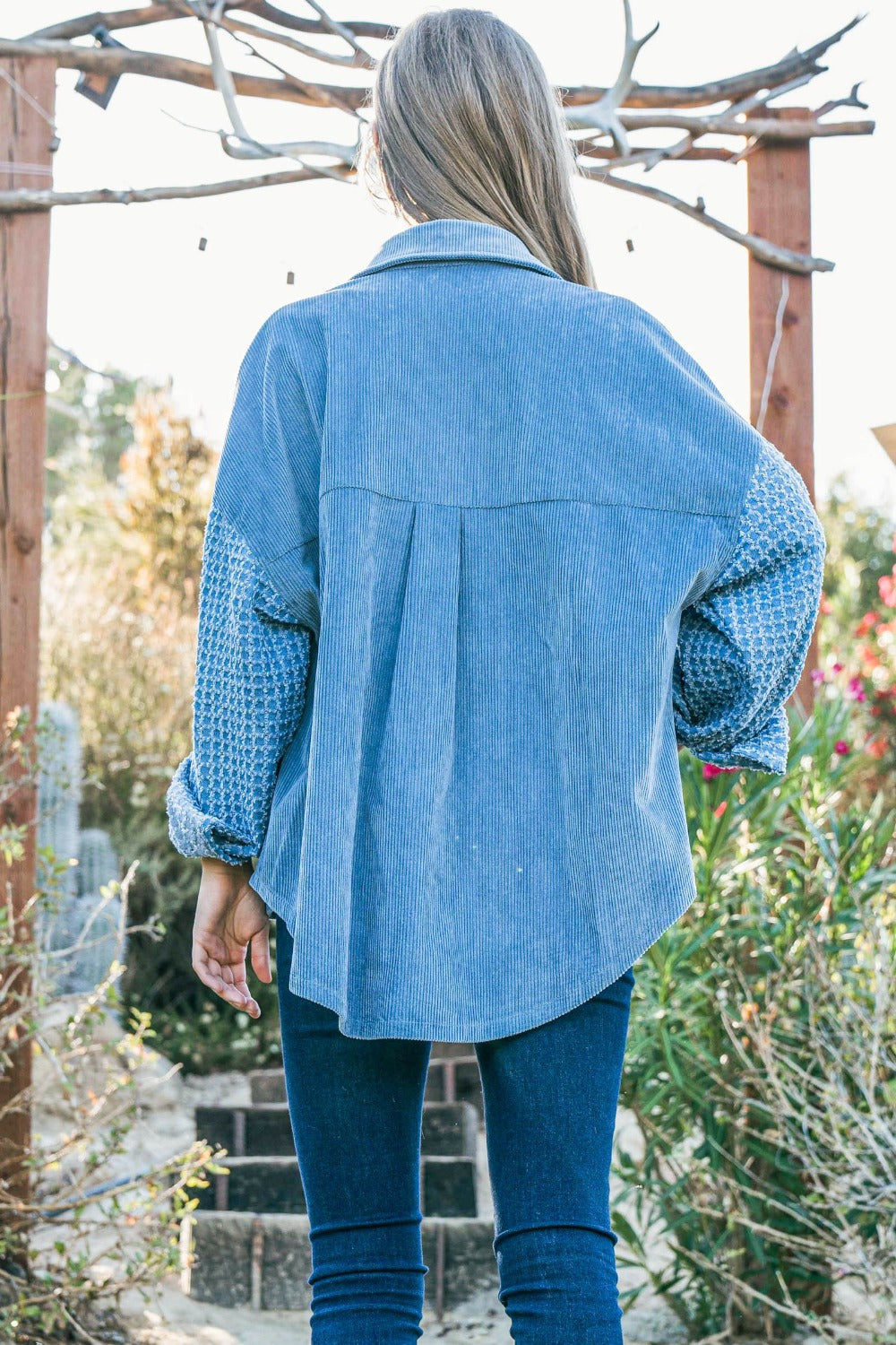 A woman stands outdoors wearing the And The Why Button Down Corduroy Contrast Texture Long Sleeve Shacket over a white top and blue jeans. She has long hair and wears a belt with a prominent buckle.