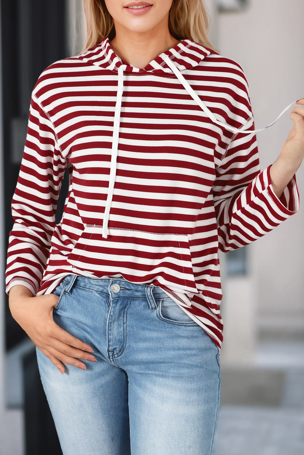 A person wearing a trendy Burgundy Stripe Kangaroo Pocket Drawstring Hoodie, paired with denim shorts and sunglasses, holds a white tumbler and a woven bag in front of a white paneled wall.