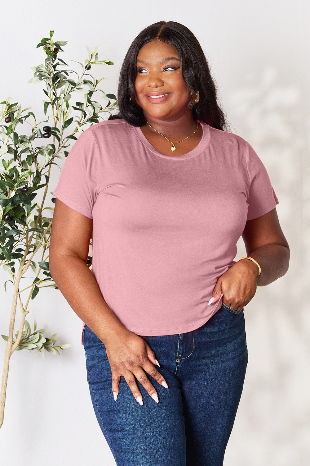 A woman stands next to an indoor plant, smiling, with one hand in her khaki pants pocket. In her pink Basic Bae Full Size Round Neck Short Sleeve T-Shirt and gold jewelry, she radiates casual elegance.