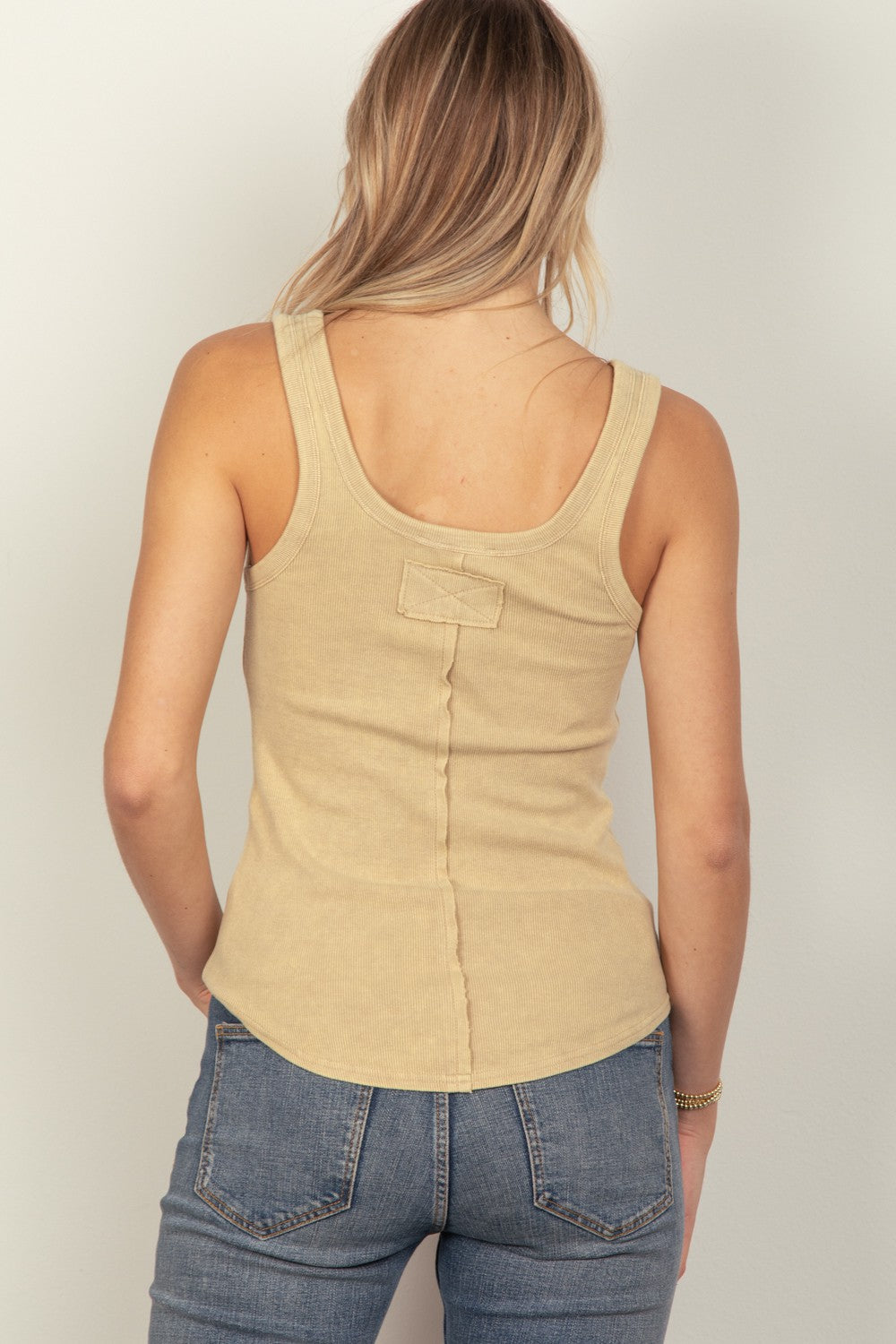 A woman with long blonde hair is wearing the VERY J Washed Ribbed Tank with Placket Detail in beige and blue jeans, standing against a plain background. Channeling 90's fashion, she's dressed in a spring wardrobe essential. She has a neutral expression and is looking slightly to the side.