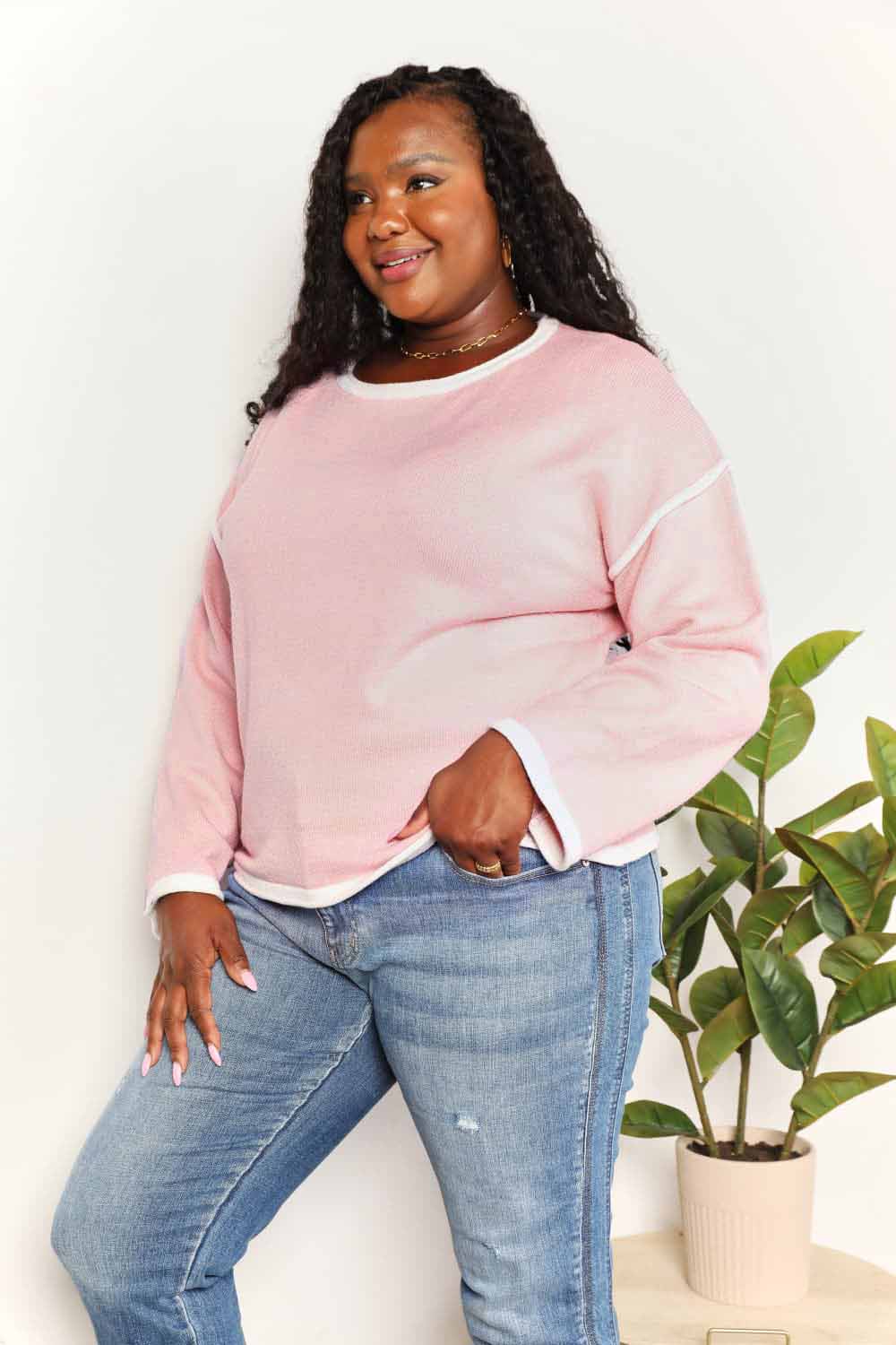 A woman with long curly hair is wearing the Angel Wings Contrast Detail Dropped Shoulder Knit Top in pink, paired with blue jeans, standing against a light background.