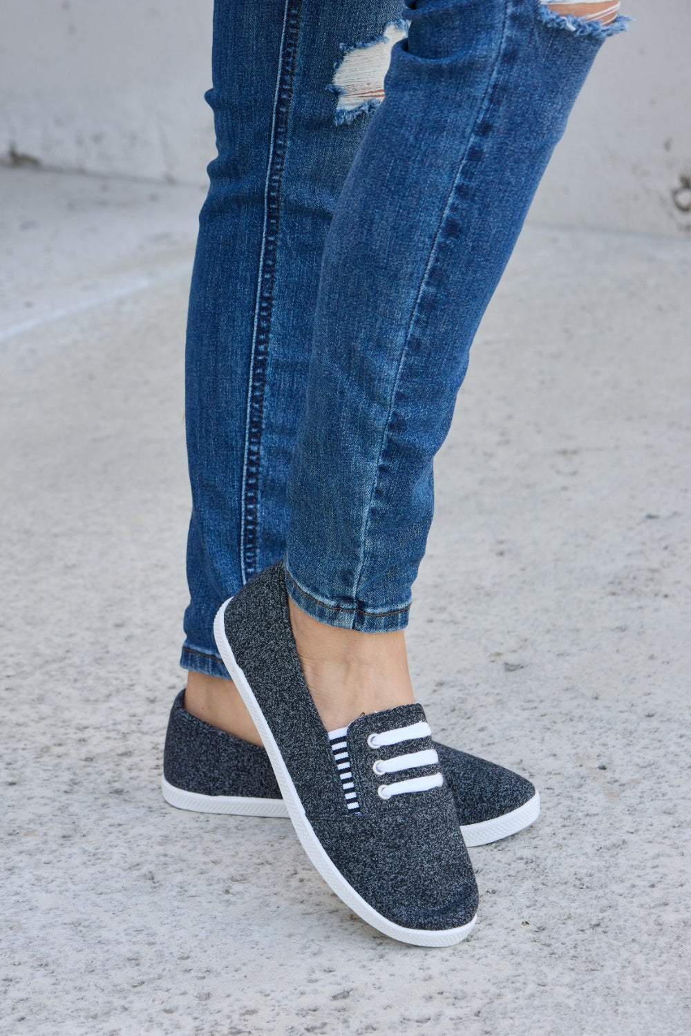 A person wearing ripped blue jeans and the fashionable Forever Link Round Toe Slip-On Flat Sneakers with white elastic bands.