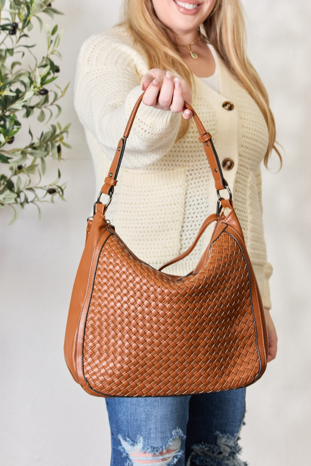 A person carries the SHOMICO Weaved Vegan Leather Handbag in a stylish brown hue, featuring a sleek crossbody strap, expertly paired with a cream sweater and jeans.