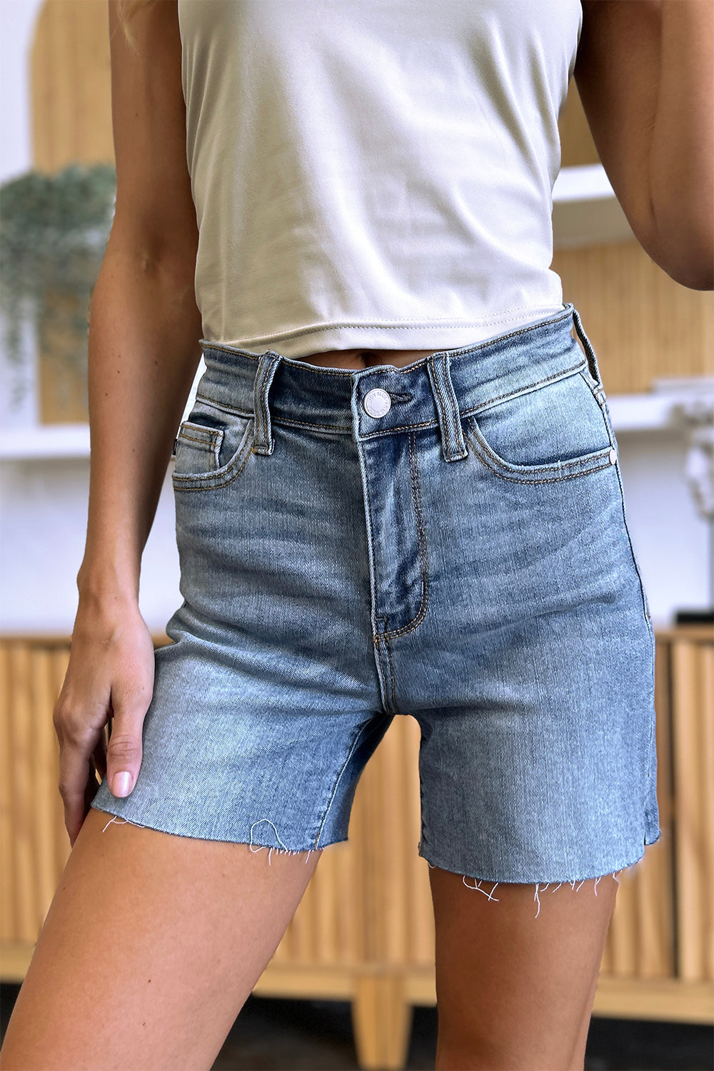 Side view of a person wearing a white shirt and Judy Blue Full Size High Waist Raw Hem Denim Shorts, standing indoors near a wooden cabinet. Summer fashion vibes radiate through the casual ensemble.