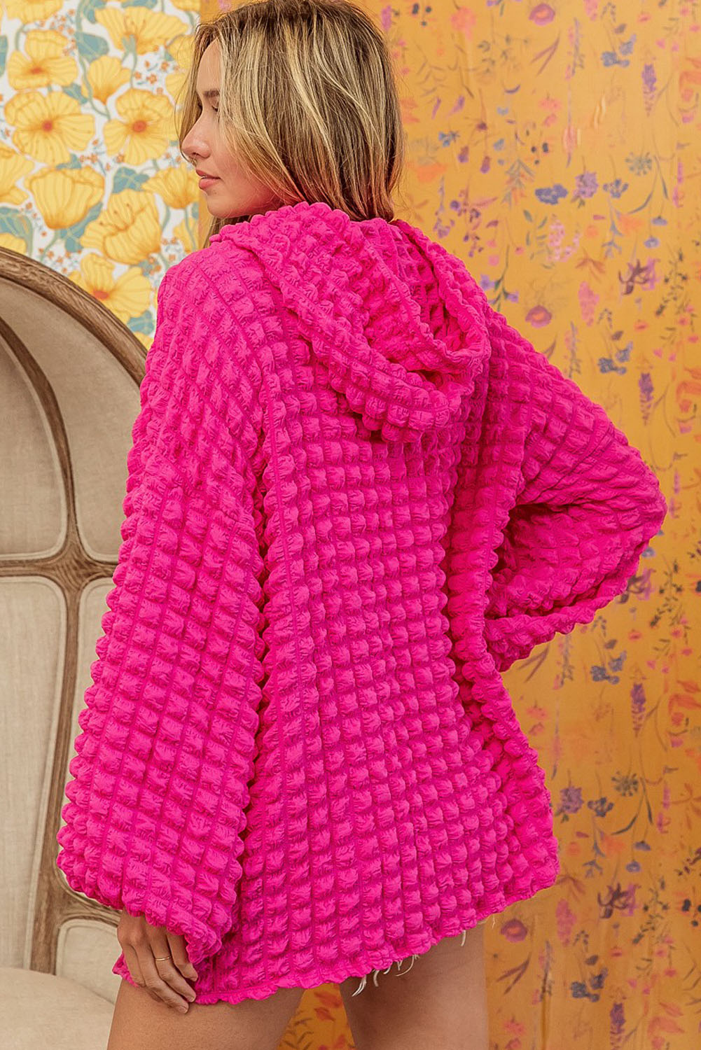 A person stands with their back to the camera, donning the Rose Bubble Textured Waffle Hoodie in a vibrant pink hue. They are in a room with yellow floral wallpaper, and a corner of a wooden chair is visible.