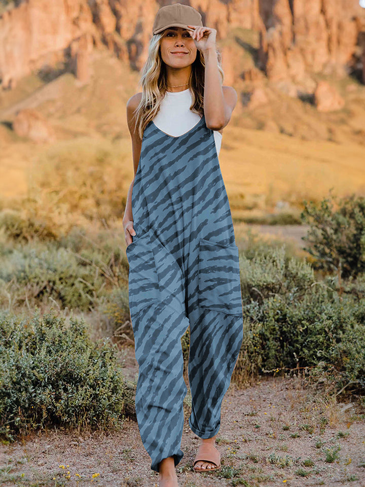 A person wearing a casual white V-neck tank top and a Full Size Printed V-Neck Sleeveless Jumpsuit with a brown animal print zebra-stripe pattern stands outdoors with one hand on their hat and the other in their pocket, set against a rocky landscape backdrop.