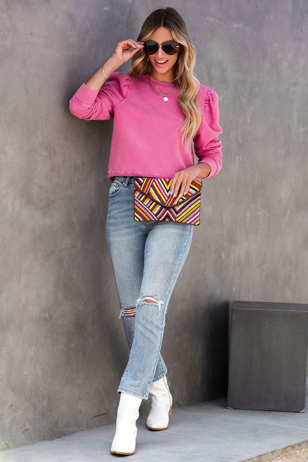 A woman with long blonde hair and a vintage look is wearing sunglasses, a Rose Vintage Washed Puff Sleeve Sweatshirt, and light blue jeans. She has one hand on her hip and is holding a colorful patterned clutch.