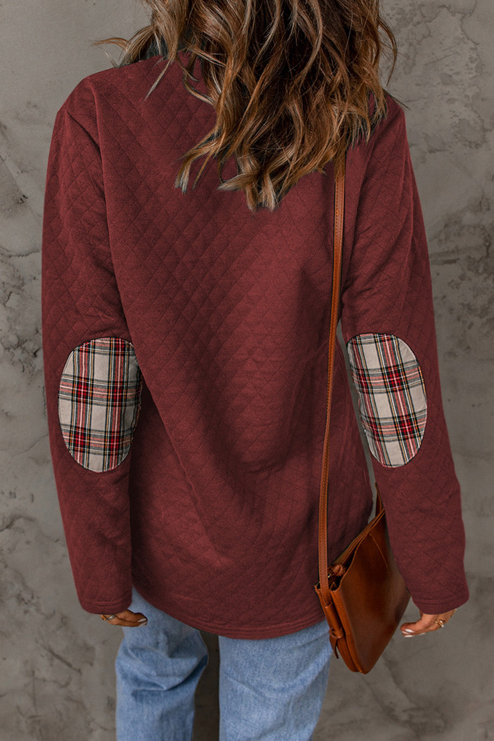 A person with wavy hair is wearing a chic Fiery Red Geometric Texture Plaid Trim Sweatshirt. They have a brown crossbody bag and are facing away from the camera, with a gray textured wall in the background.