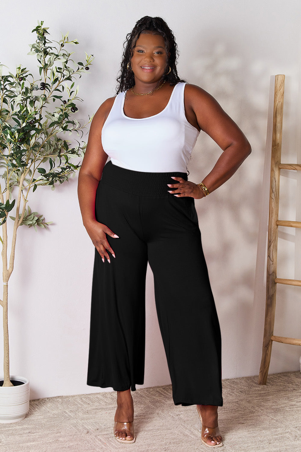 A curve model is wearing the Double Take Full Size Smocked Wide Waistband Wide Leg Pants in gray with hands in pockets, paired with white sneakers.