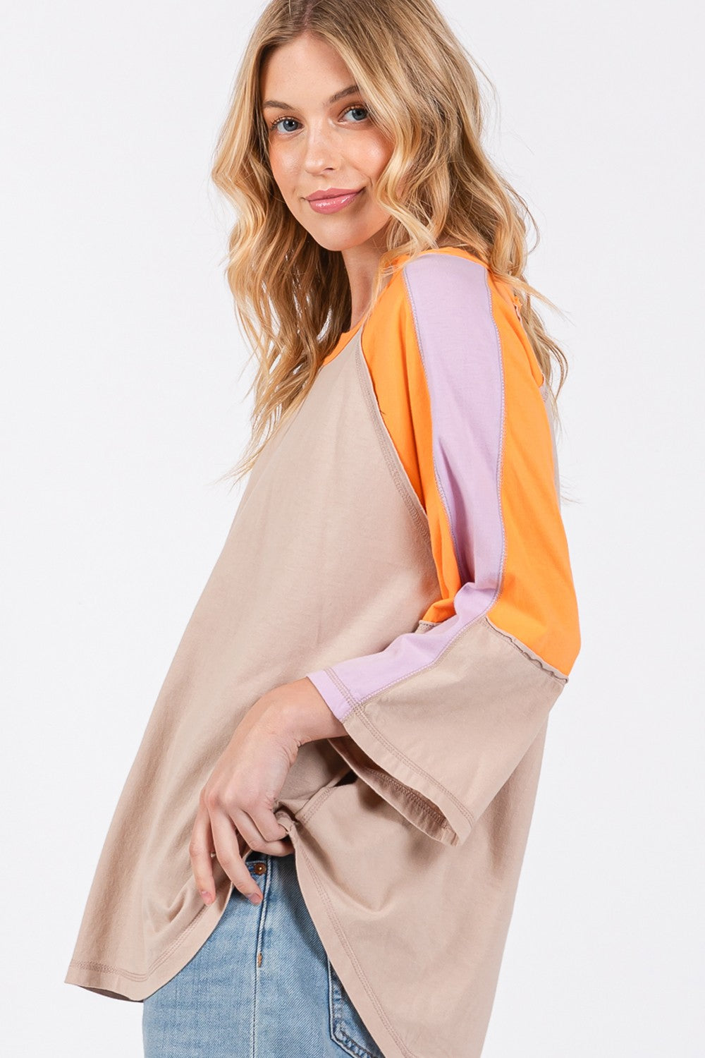 Person wearing a chic modern look with the SAGE + FIG Color Block Curved Hem T-Shirt in beige, featuring orange and purple accents, paired with blue jeans and a hat, poses against a plain background.