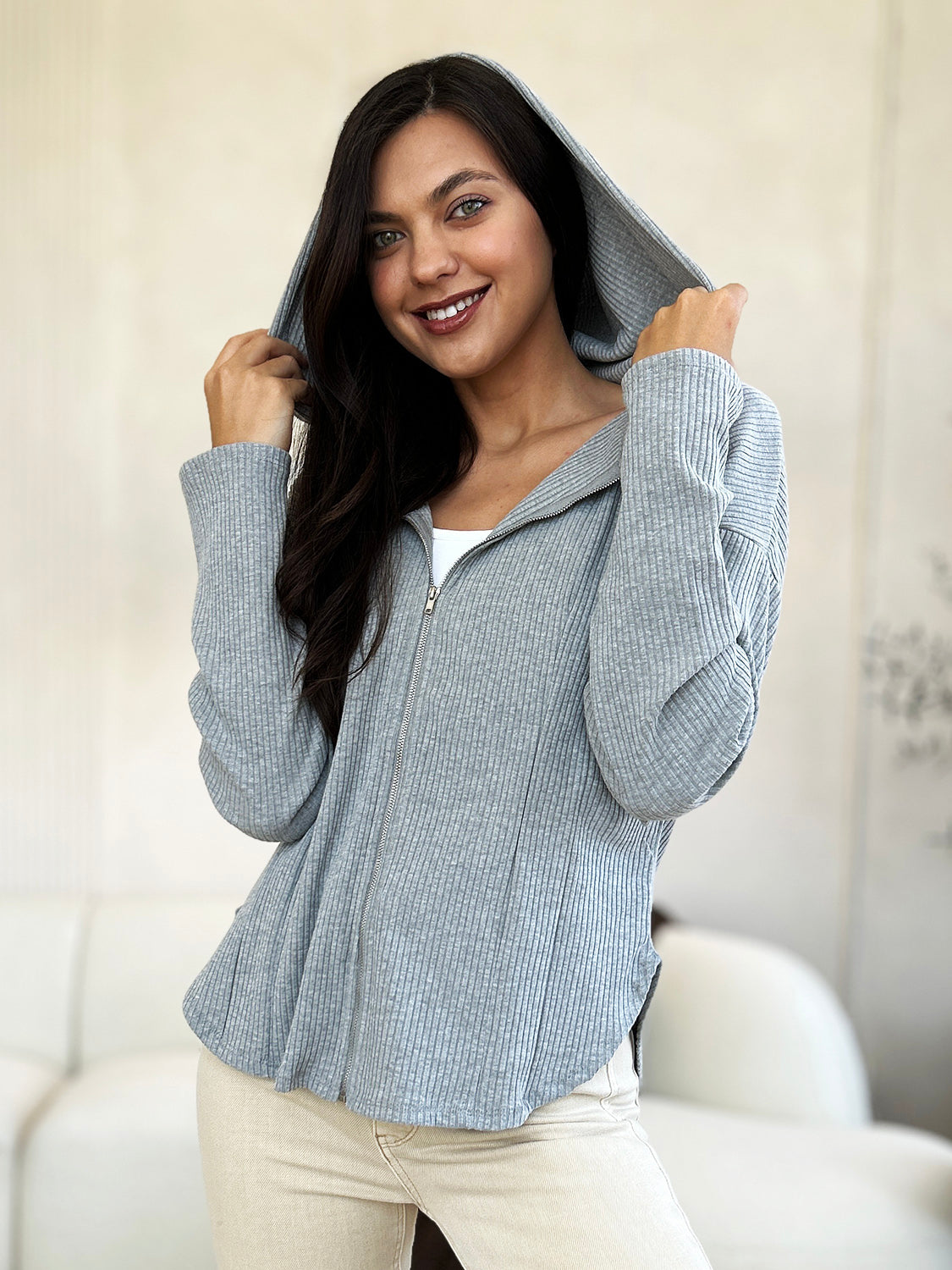 A woman in a trendy Culture Code Full Size Ribbed Zip Up Drawstring Hooded Jacket and light-colored pants stands indoors, smiling with her eyes closed and one hand resting on her chin.