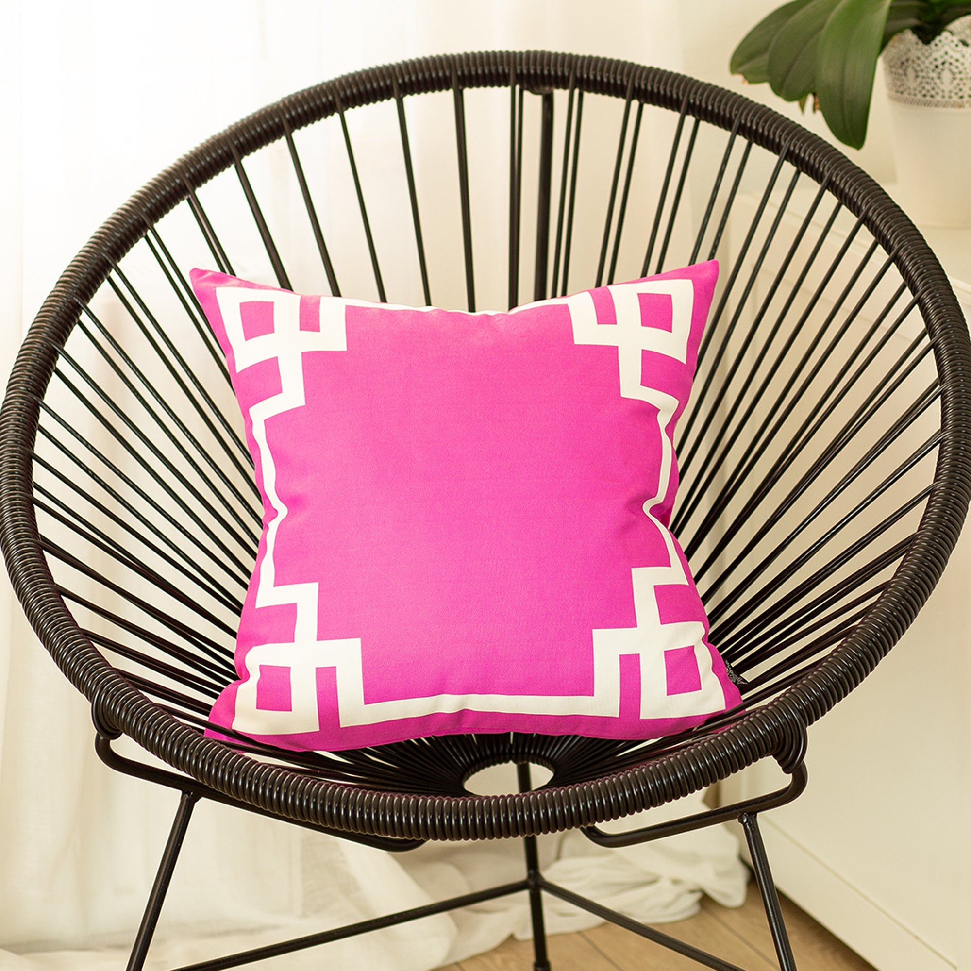 A round black wire chair, adorned with a Geometric Pink&White Square 18" Throw Pillow Cover & Insert, featuring high-quality digital printing, sits indoors on a wooden floor near a plant.