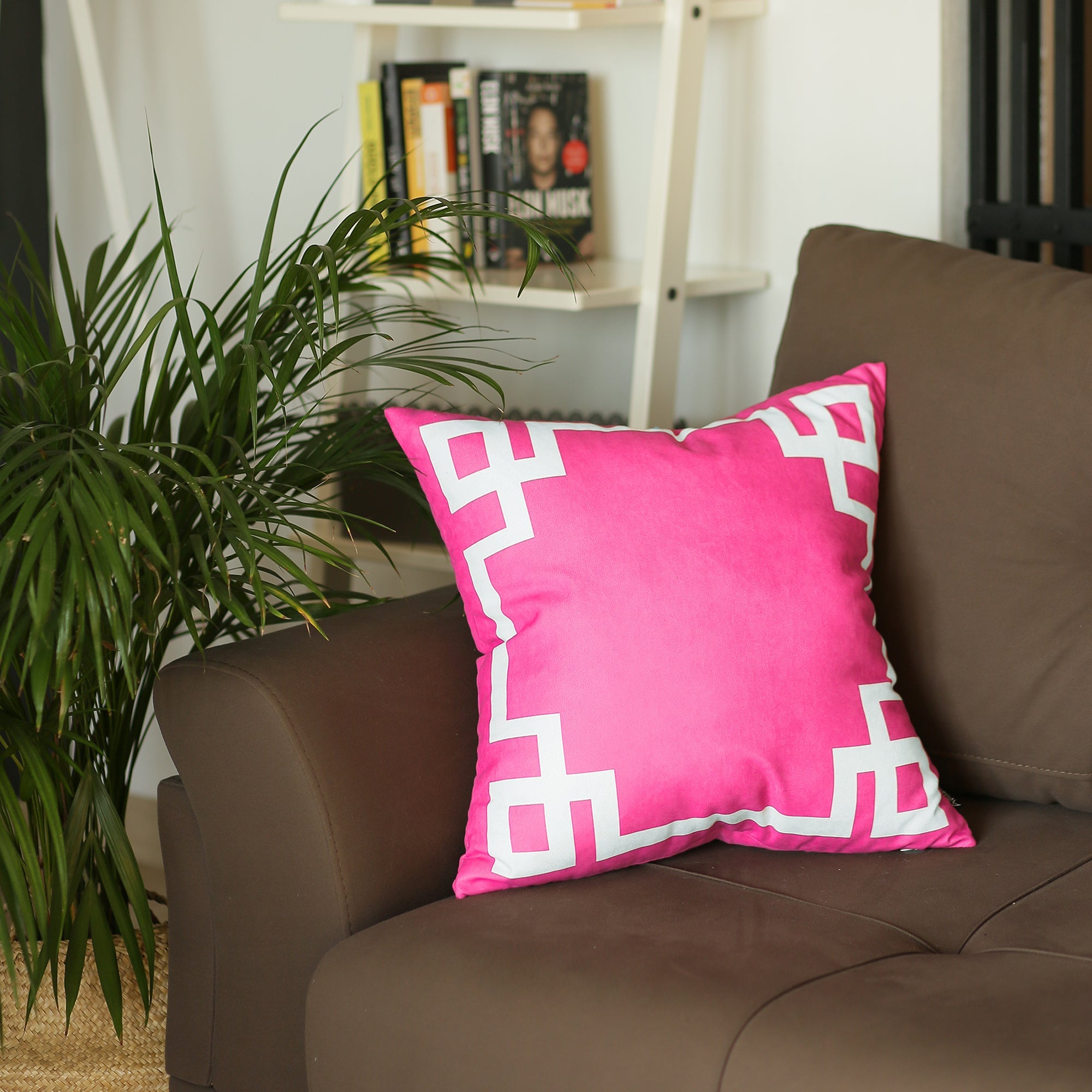 A round black wire chair, adorned with a Geometric Pink&White Square 18" Throw Pillow Cover & Insert, featuring high-quality digital printing, sits indoors on a wooden floor near a plant.