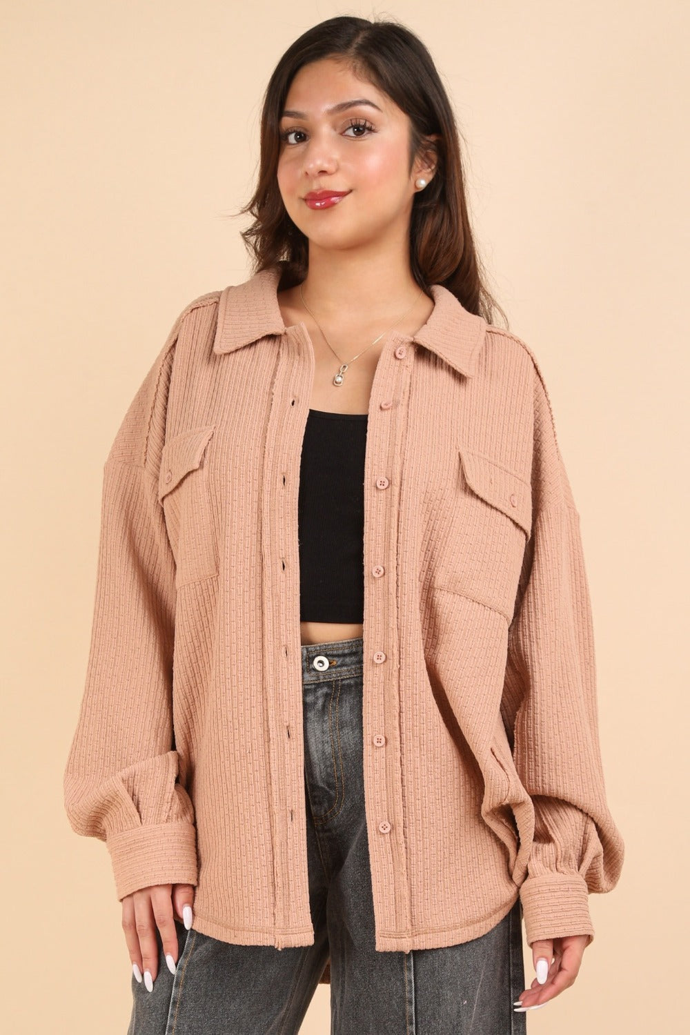 A woman wearing the VERY J Button Down Textured Knit Shacket in an oversized beige style over a black top stands against a plain background, showcasing the perfect transitional piece for any wardrobe.