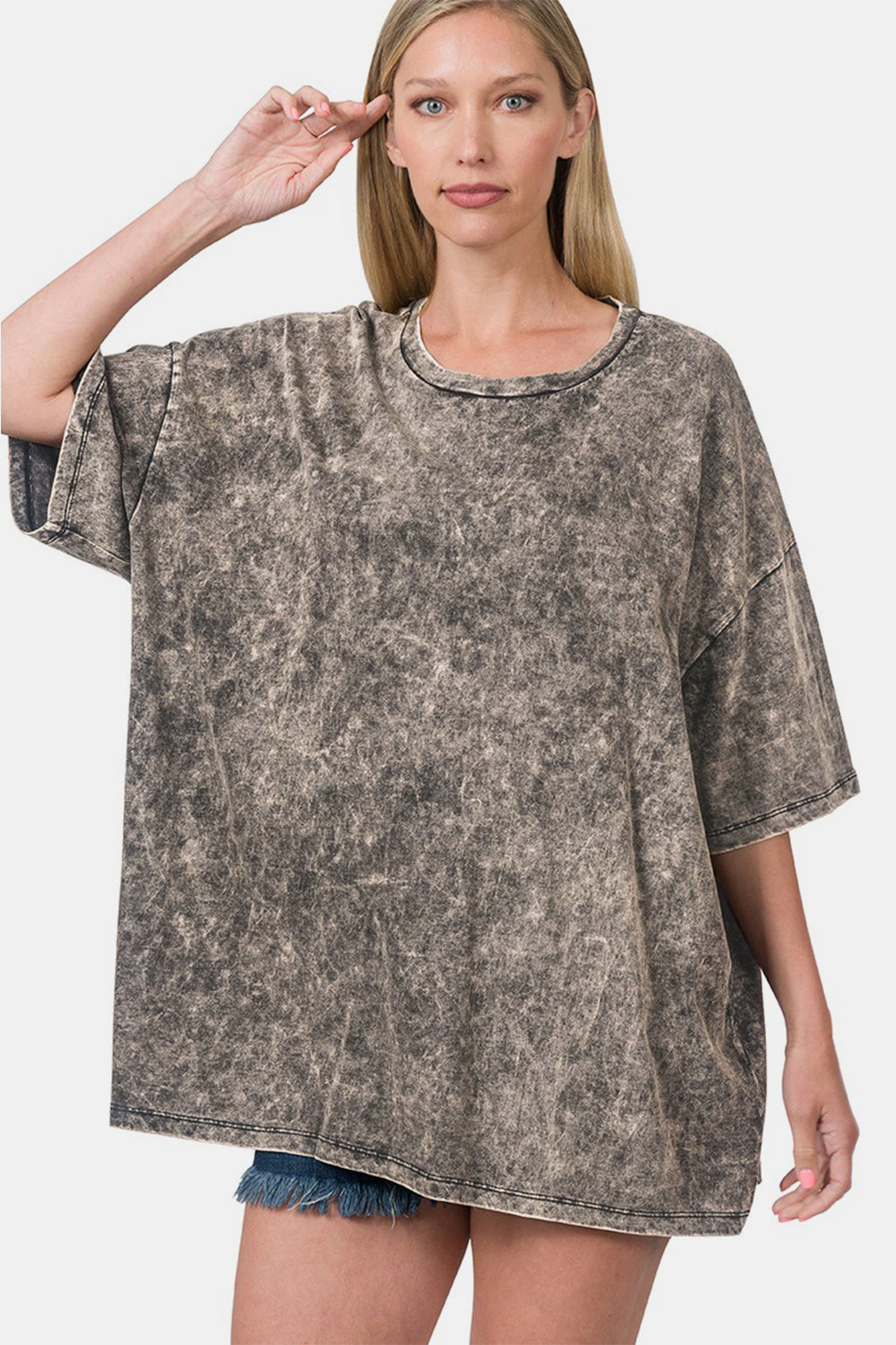 A woman is wearing a Zenana Washed Round Neck Drop Shoulder Oversized T-Shirt paired with denim shorts. Adding a trendy twist, she stands with one hand touching the brim of the t-shirt.