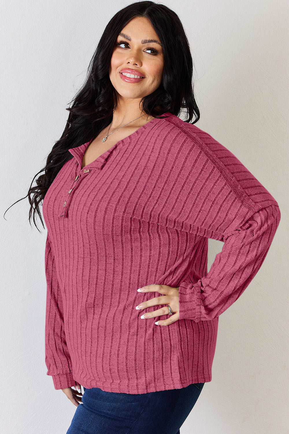 A woman with long, wavy blonde hair wears a slightly stretchy, textured Basic Bae Full Size Ribbed Half Button Long Sleeve T-Shirt in pink, standing against a neutral backdrop.